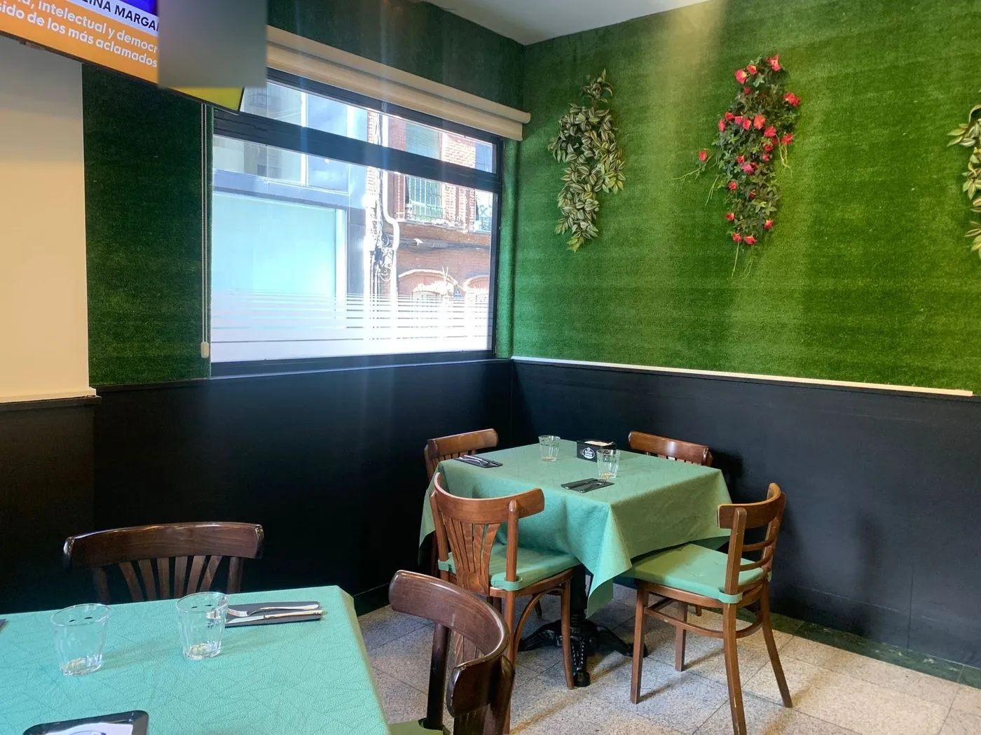 Rincón del comedor con pared de césped artificial, flores colgadas y mesas con mantel verde