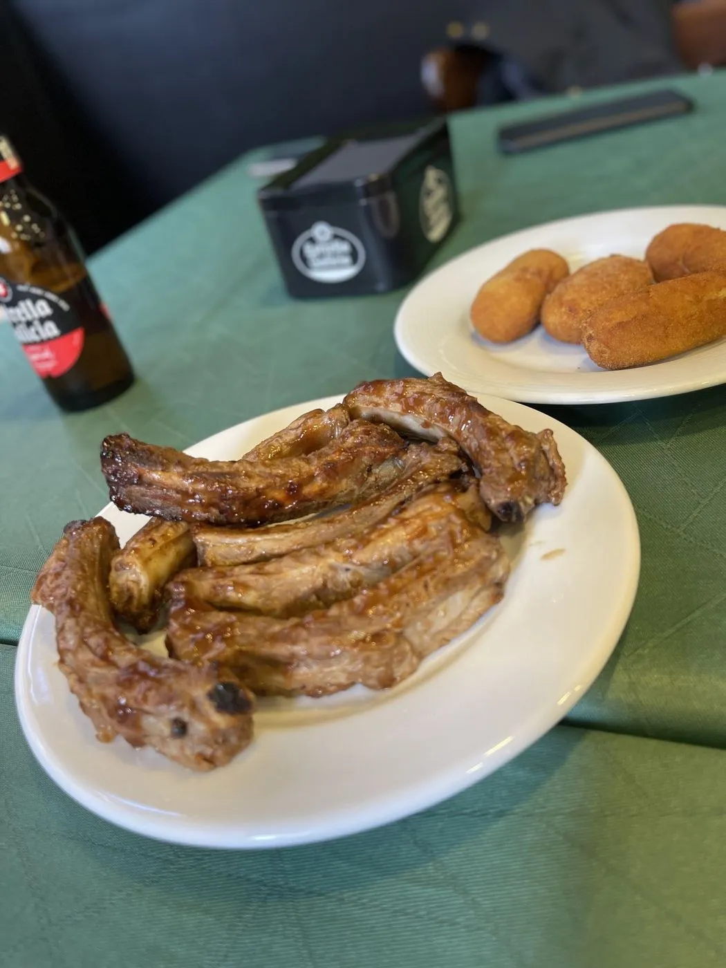 Plato de costillas glaseadas junto a otro de croquetas sobre un mantel verde